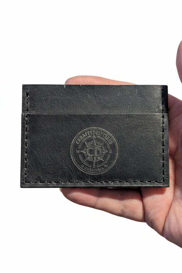 Handmade black leather card holder with embossed compass logo held in a hand.