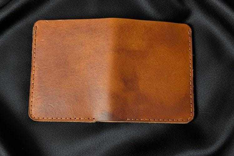 Handmade tan leather bifold wallet with visible stitching on a black fabric background.