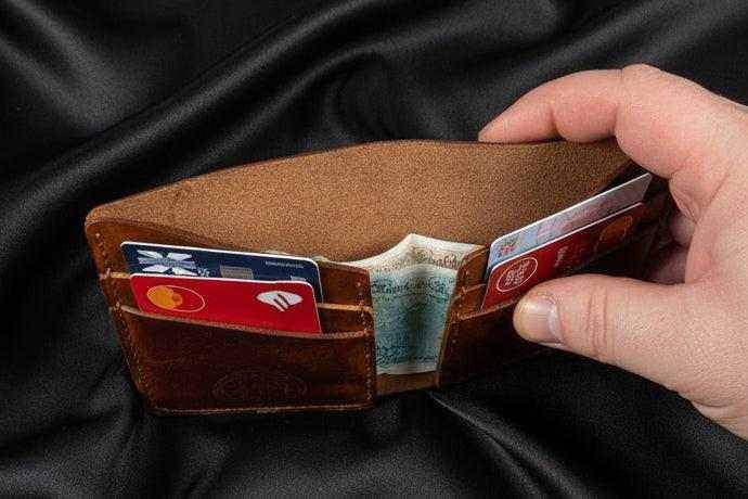 Handmade brown leather wallet open showing credit cards and cash on black fabric.