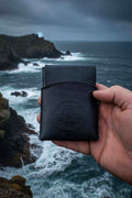 Hand holding a handmade black leather wallet with an embossed logo against a rocky Cornish coast.