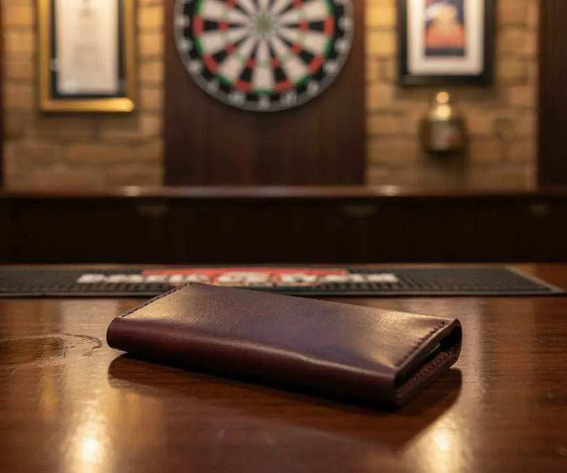 Tri-Fold brown leather darts case closed on a pub table in front of a dartboard