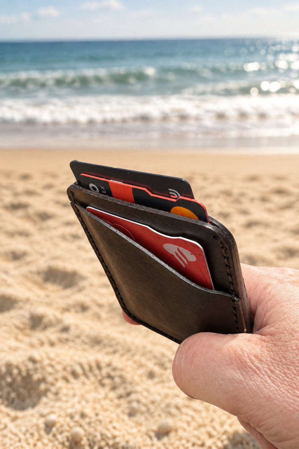 Dark Badalassi leather card holder held in hand on a beach, cards fanned inside