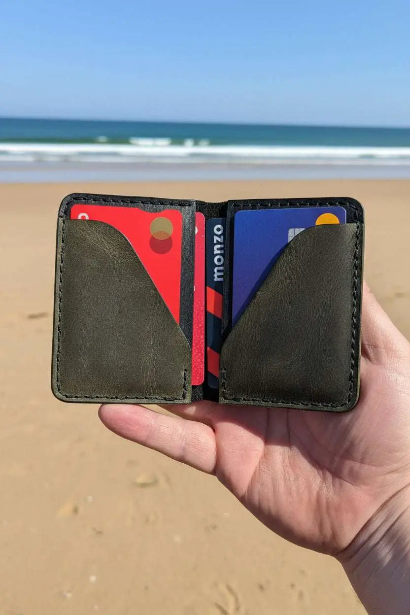 Handmade olive green leather bifold wallet with credit cards, held against a beach backdrop.