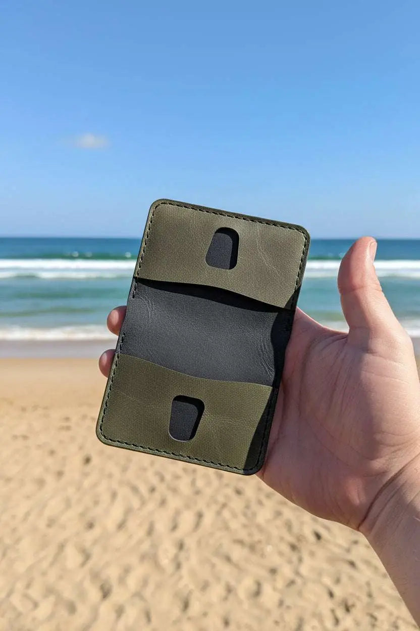 Hand holding an open olive green handmade leather wallet on a sandy beach with ocean background