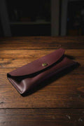Handmade burgundy leather glasses case with brass snap closure on a rustic wooden surface