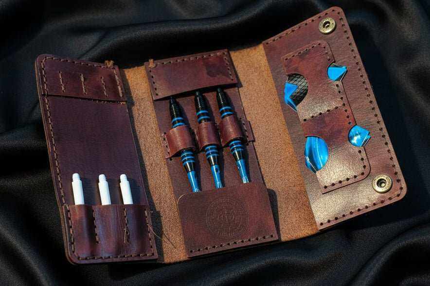 Tri-Fold leather darts case open on dark fabric, holding darts, flights and shafts